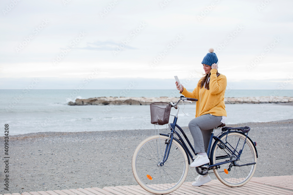 Fototapeta premium Beautiful active mature woman with bicycle on the road by the sea. Happy smiling middle aged woman with headphones listening to music, copy space