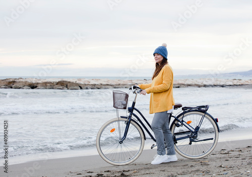 brunette woman in hat in spring with her bicycle walking the coast by the sea beach.