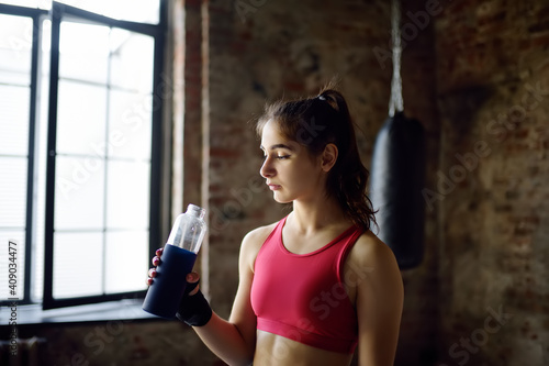 Portrait of young beautiful athlete in a boxing gym. A boxer girl drinks beverage from a bottle during a workout. In the process of training need to drink a lot water.