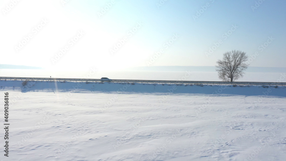 Aerial shot of white car riding through snow covered road near a frozen lake. SUV driving at countryside route near icy pond at sunny winter day. Flying over the auto moving through scenic landscape
