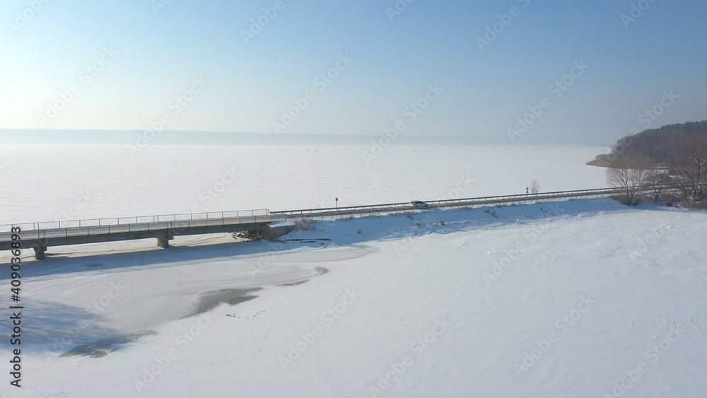 Aerial shot of white car riding through snow covered road near a frozen lake. SUV driving at countryside route near icy pond at sunny winter day. Flying over the auto moving through scenic landscape
