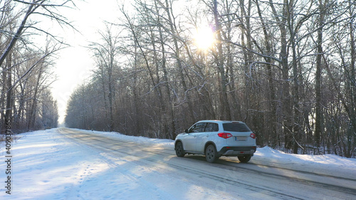 White car fast riding through snow covered icy road. SUV going at empty countryside route in winter forest on sunny day. Auto moving through scenic landscape way. Travel concept. Top view Drone shot