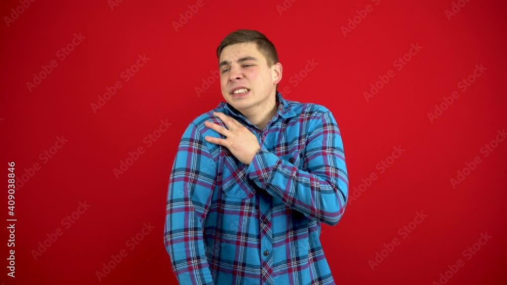 A young man has a sore shoulder and he touches it with his hand. Shooting on a red background. A young man has a sore shoulder and he touches it with his hand. Shooting on a red background.