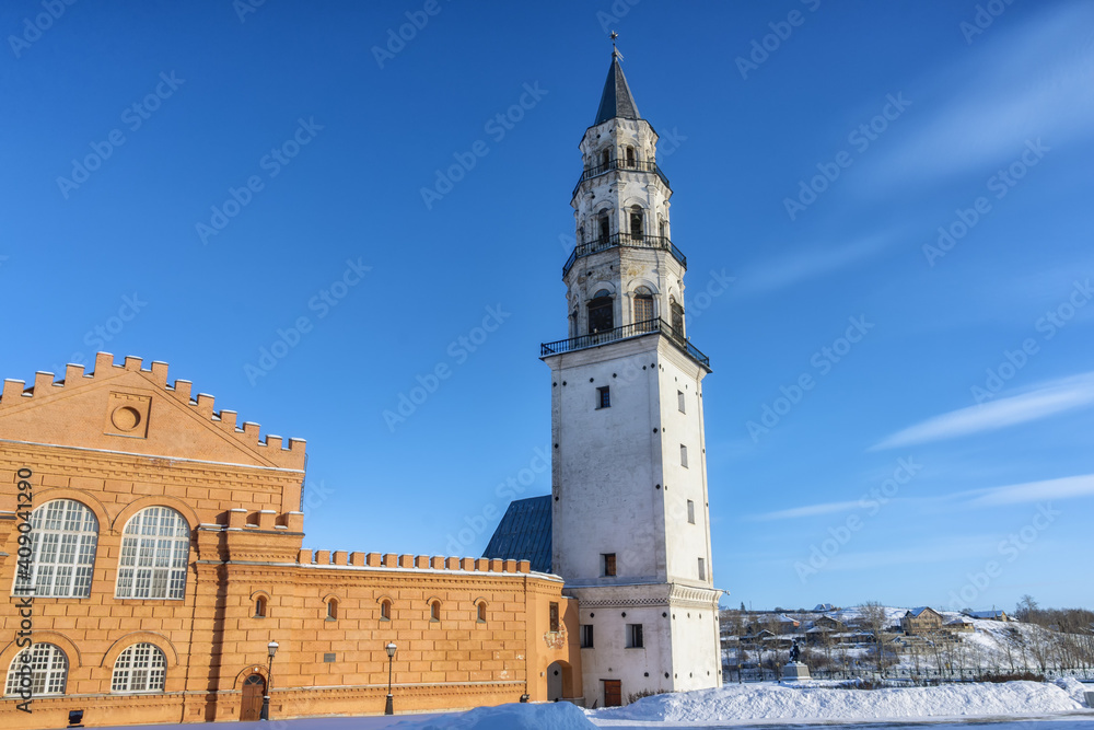 An inclined white-stone multi-storey tower in the provincial town of Nevyansk (Russia) on a winter day against the backdrop of a blue sky. A city with wooden houses is visible in the distance 