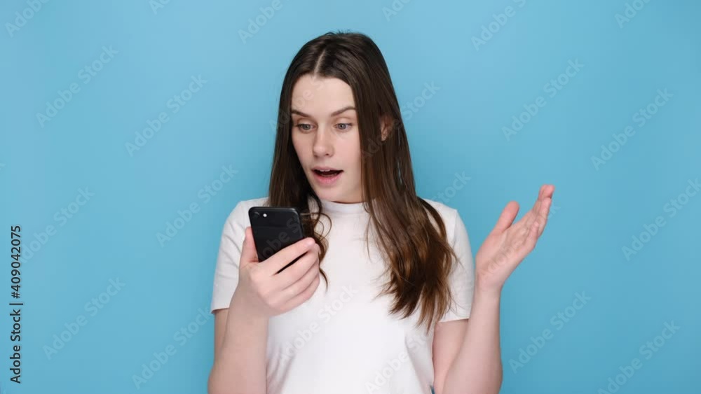 Surprised cute woman points at smart phone device, suggests to have conversation in group chat, glad to recieve many messages of congratulation on mailbox, isolated on blue wall, has overjoyed look Surprised cute woman points at smart phone device, suggests to have conversation in group chat, glad to recieve many messages of congratulation on mailbox, isolated on blue wall, has overjoyed look