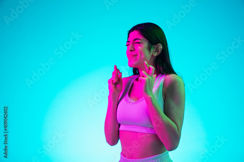 Fingers crossed, luck. Brunette woman's portrait isolated on blue studio background in mixed neon. Beautiful model in white top. Concept of human emotions, facial expression, sales, ad, fashion.