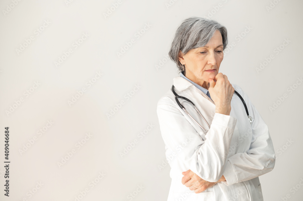 Portrait of a senior mature female doctor standing with a stethoscope, posing over gray background, looking down and thinking, skilled and experienced healthcare professional wearing a medical robe Portrait of a senior mature female doctor standing with a stethoscope, posing over gray background, looking down and thinking, skilled and experienced healthcare professional wearing a medical robe