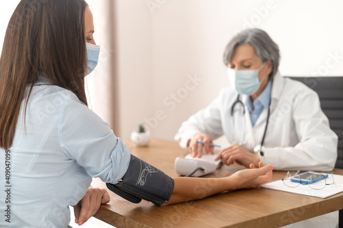 Senior mature female doctor wearing a lab coat and a face mask  using sphygmomanometer with stethoscope checking blood pressure to a sick patient in the hospital at the doctor office during pandemic
