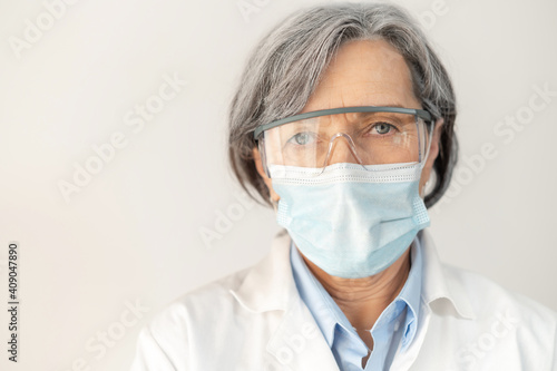 A close-up of a senior gray-haired female doctor wearing protective goggles, a face mask, and a medical robe, portrait of a fully equipped middle-aged healthcare professional working during pandemic