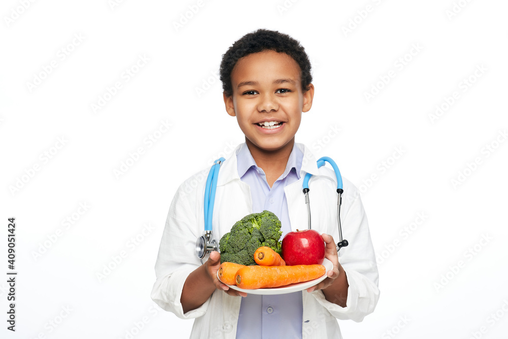 Young African American child, dressed as a doctor, recommends vegetables and fruits for proper nutrition and benefits to your health Young African American child, dressed as a doctor, recommends vegetables and fruits for proper nutrition and benefits to your health