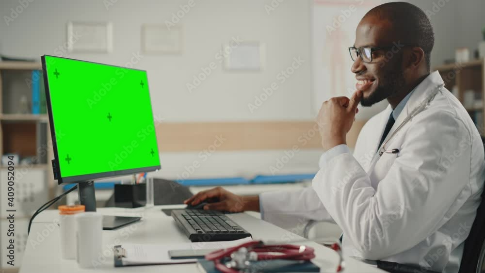Experienced African American Male Doctor Wearing White Coat Working on Computer with Green Screen Mock Up Display. Medical Health Care Professional Working with Test Results and Patient Treatment. Experienced African American Male Doctor Wearing White Coat Working on Computer with Green Screen Mock Up Display. Medical Health Care Professional Working with Test Results and Patient Treatment.