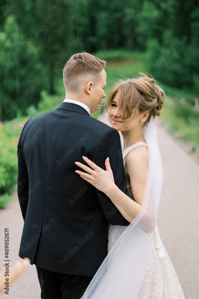 Smiling young woman in wedding dress posing on camera with handsome husband standing behind and holding hands on her waist.