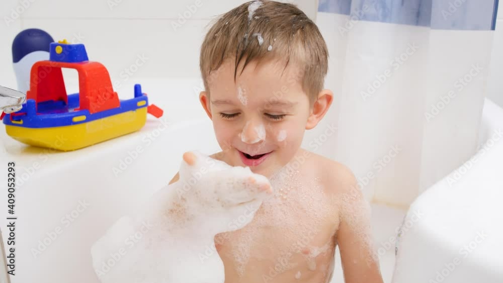 Slow motion of cheerful laughing boy having bath and blowing soap foam from hand in camera. Concept of child hygiene and health care at home. Kids having fun and playing Slow motion of cheerful laughing boy having bath and blowing soap foam from hand in camera. Concept of child hygiene and health care at home. Kids having fun and playing