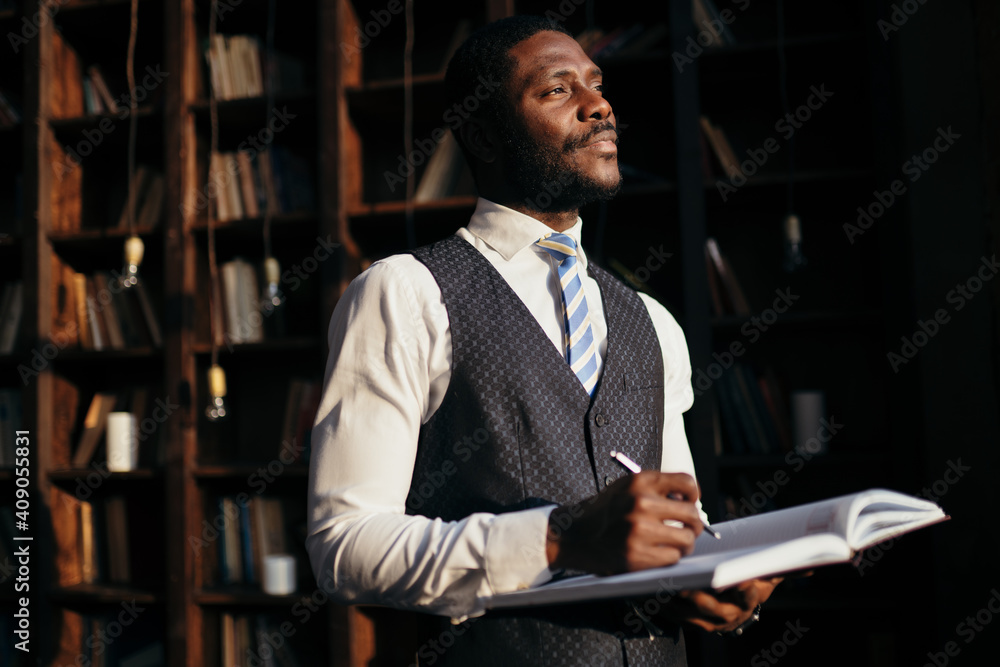 African american man holding a notebook with a pen in his hands among the shelves with books