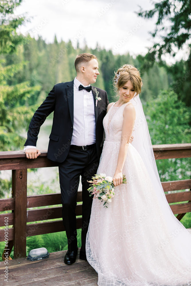 Full length portrait of handsome man in stylish black tuxedo suit embracing his charming young bride in white wedding dress. Happy newlywed couple posing near forest lake. Full length portrait of handsome man in stylish black tuxedo suit embracing his charming young bride in white wedding dress. Happy newlywed couple posing near forest lake.