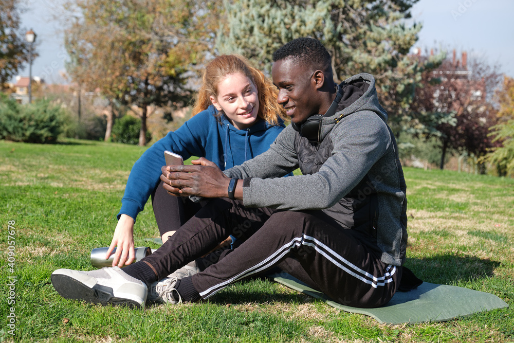 African black man and redhead caucasian woman checking the smartphone after exercising in a park with a mat. Fitness and sport outdoor. African black man and redhead caucasian woman checking the smartphone after exercising in a park with a mat. Fitness and sport outdoor.