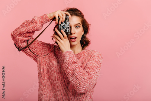 Surprised girl in knitted sweater taking photos. Studio shot of female photographer holding camera on pink background.