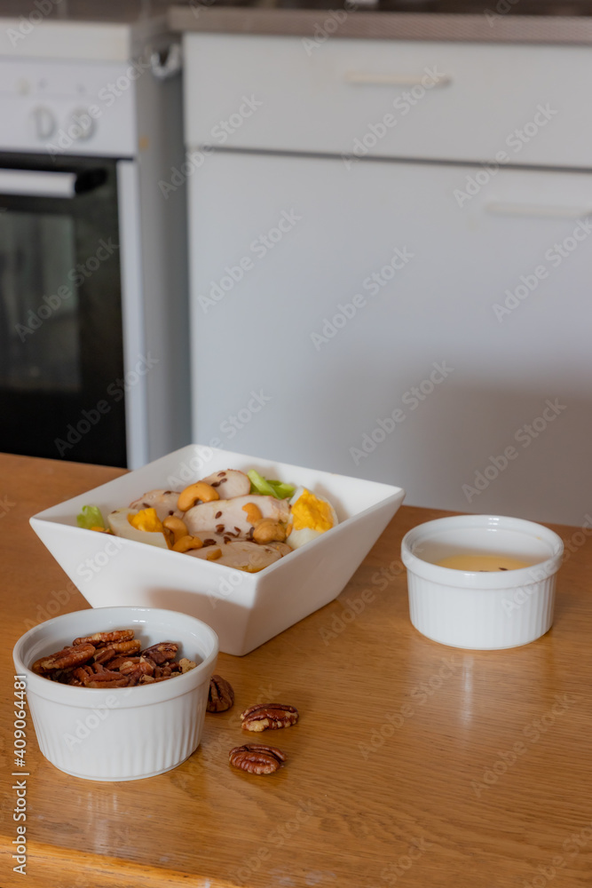 Chicken salad with lettuce, egg, peanuts and flaxseed in a white square bowl, small bowls one with dressing and one with walnuts on a wooden table, blurred background. Healthy and nutritious food Chicken salad with lettuce, egg, peanuts and flaxseed in a white square bowl, small bowls one with dressing and one with walnuts on a wooden table, blurred background. Healthy and nutritious food