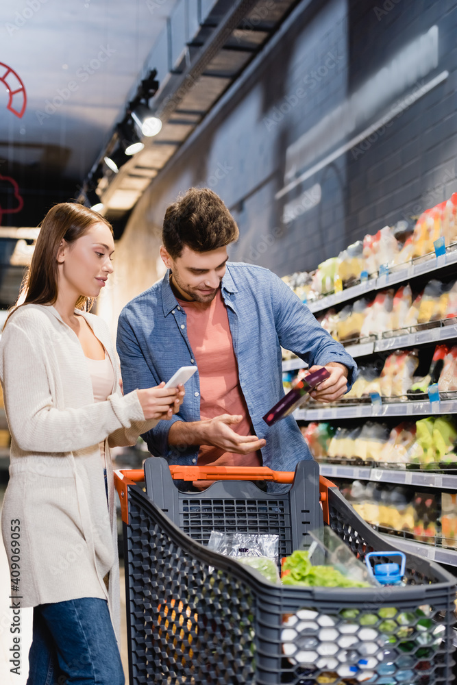 Woman holding smartphone near boyfriend with grocery and shopping cart on blurred foreground