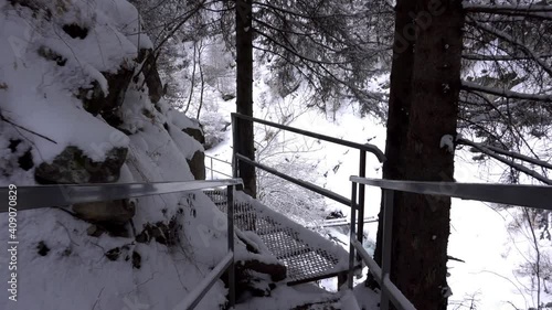 First-person view of a metal bridge in the forest. I walk across a bridge in a snowy gorge, among white rocks and trees. A clear river runs. Mountainous terrain. Snow is falling. Almarasan, Almaty photo