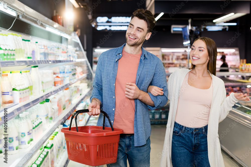Smiling couple with shopping basket looking at food on blurred foreground in supermarket