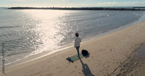 mujer madura haciendo postura loto en la playa frente al sol y dron a su espalda photo