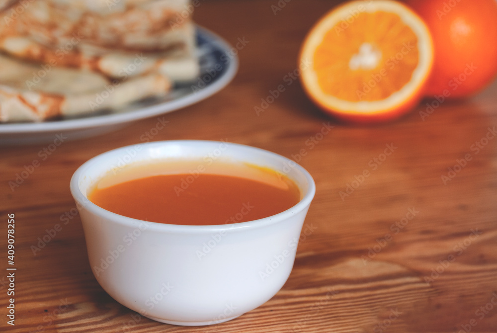 Orange syrup in a white bowl and oranges and pancakes in the background. Orange syrup in a white bowl and oranges and pancakes in the background.