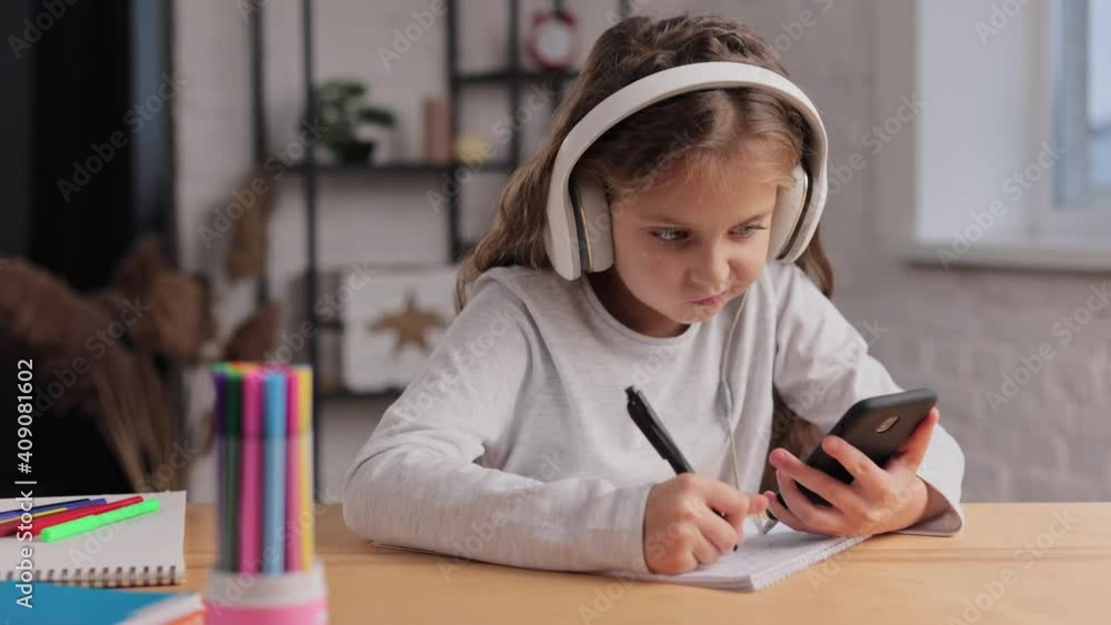 Cute primary school girl in earphones studying at home using smartphone. Schoolgirl has online lesson, writing in exercise book, doing homework. Distance learning online. Children home education Cute primary school girl in earphones studying at home using smartphone. Schoolgirl has online lesson, writing in exercise book, doing homework. Distance learning online. Children home education