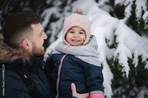 Adorable little daughter with her lovely dad stand by the big snowy tree. Cute little girl laughs