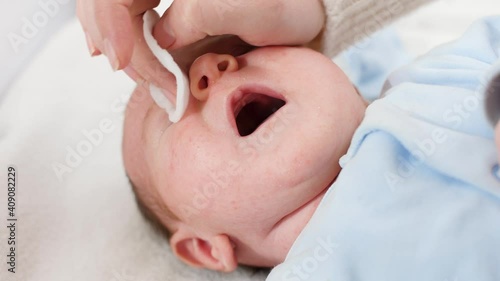 Closeup of little newbron baby boy lying in crib while mother cleaning and washig his face and eyes with cotton pad. Concept of babies and newborn hygiene and healthcare. Caring parents with little photo