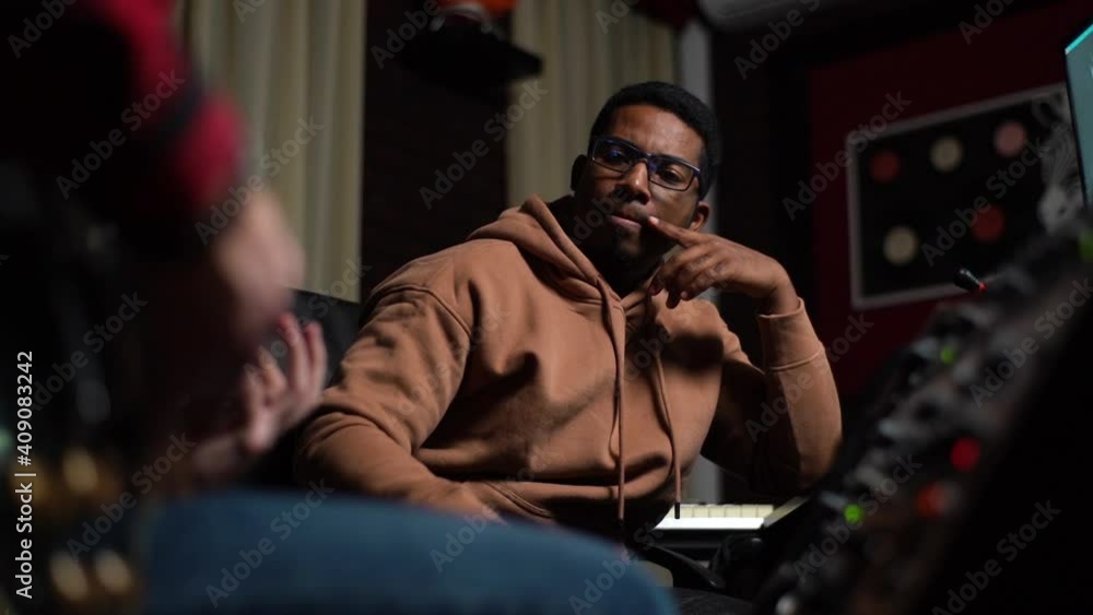 Afro-American sound producer carefully watching musician play guitar. Sound engineer looking at monitor and adjusting sound on audio mixing console, back view. Shooting in slow motion. Afro-American sound producer carefully watching musician play guitar. Sound engineer looking at monitor and adjusting sound on audio mixing console, back view. Shooting in slow motion.