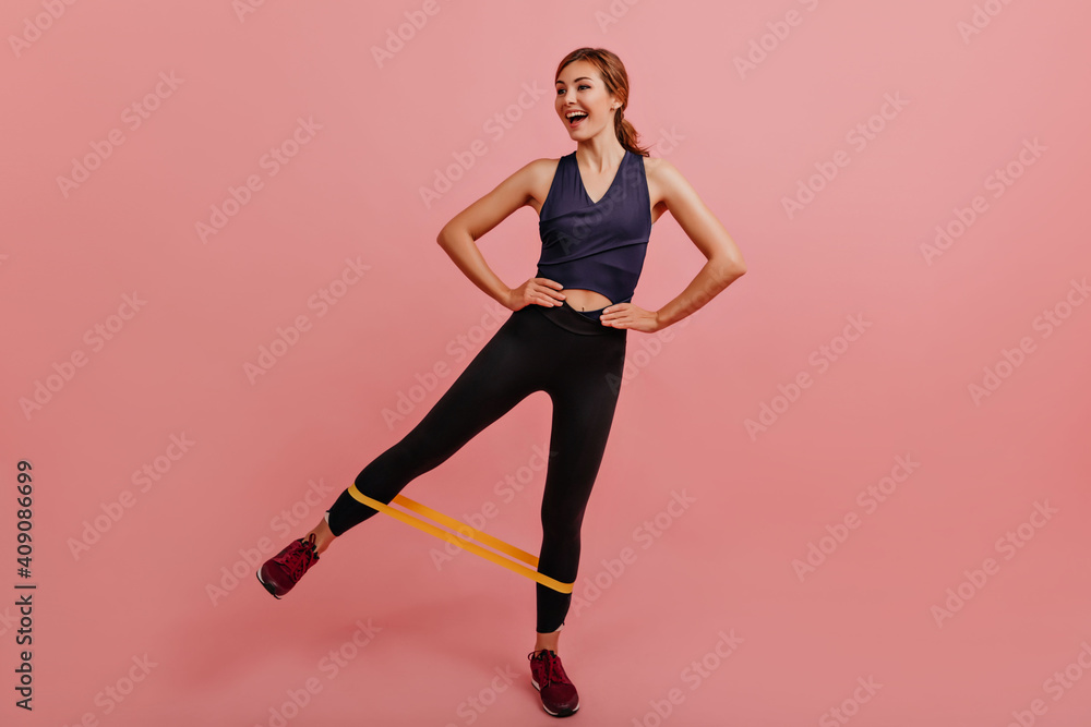 Joyful girl trains her legs with help of fitness yellow rubber bands. Full-length shot of young woman in great mood on pink background