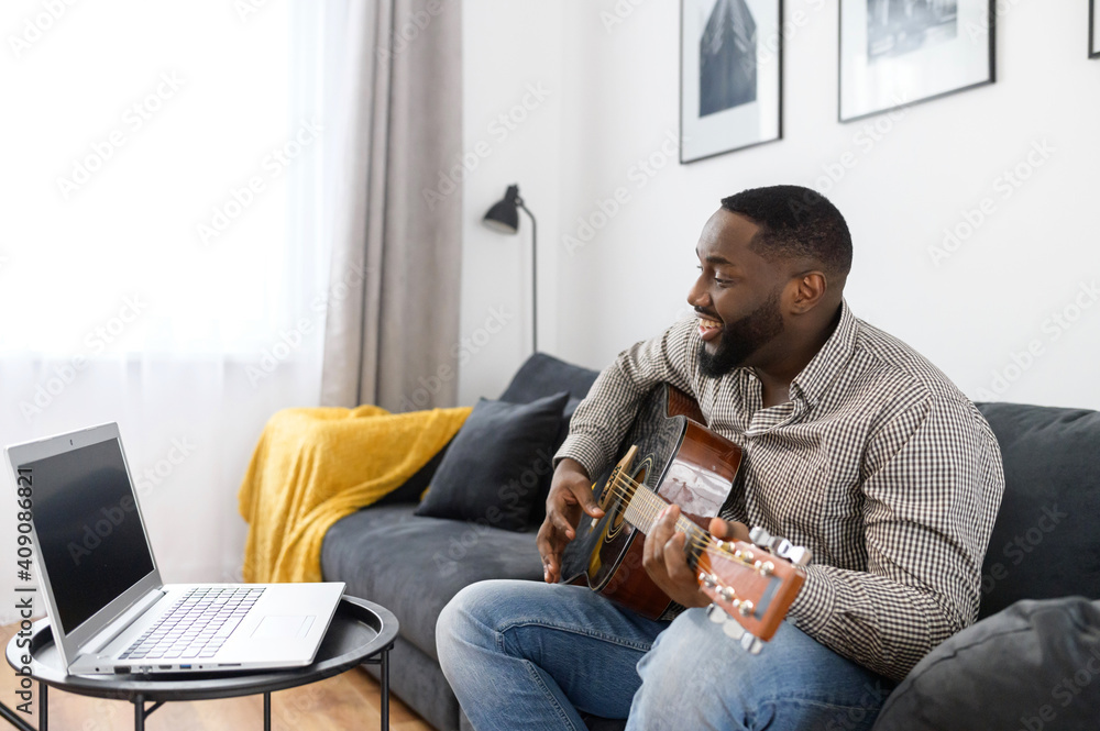 Smiling young African American male musician playing guitar during an online concert at home while isolated during coronavirus, sitting on the sofa, using a laptop to stream record or teach music - obrazy, fototapety, plakaty 