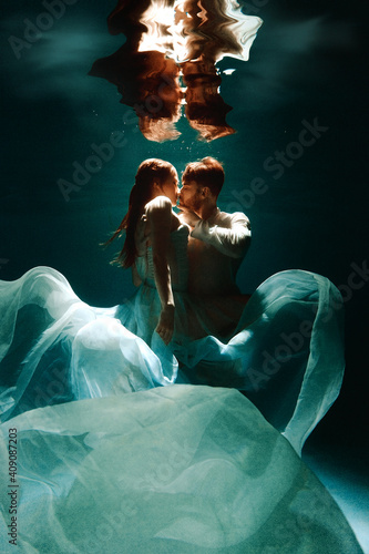The couple is dancing or hugging in the pool underwater. A girl in a dress with a long train and a guy swim underwater