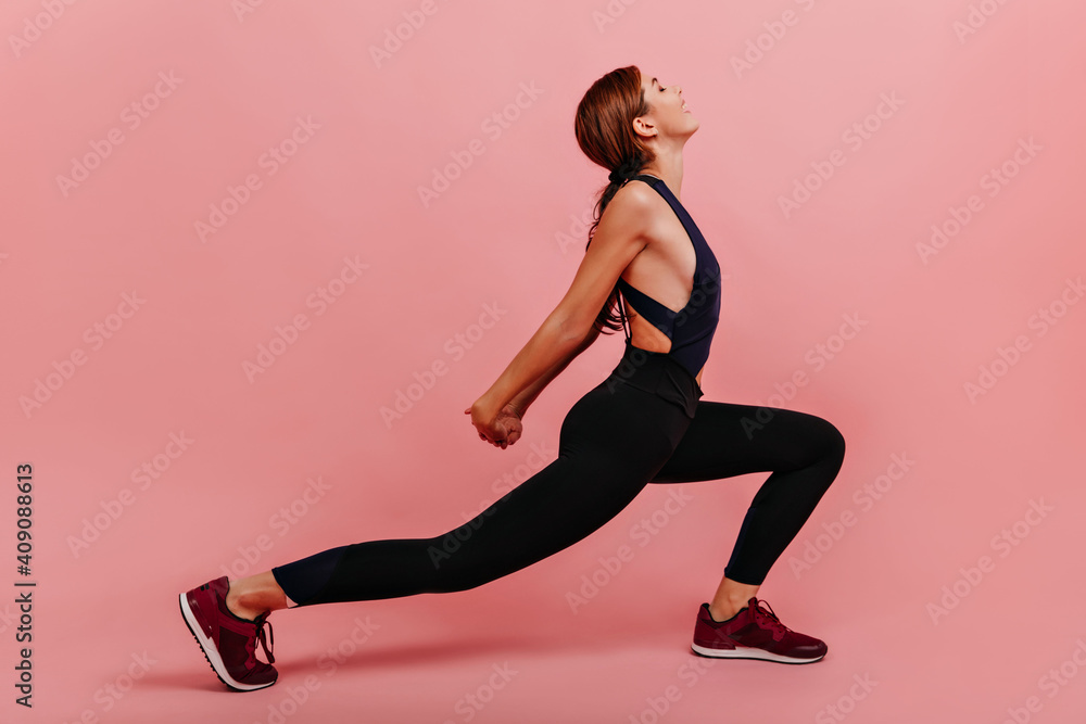 Calm athletic girl in black leggings does stretching on pink background. Photo indoor of sportswoman with ponytail doing exercise for back