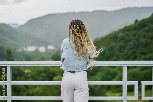 back side of a young woman with blonde braided hair wearing a denim jacket and white jean resting on a dam on a rainy day