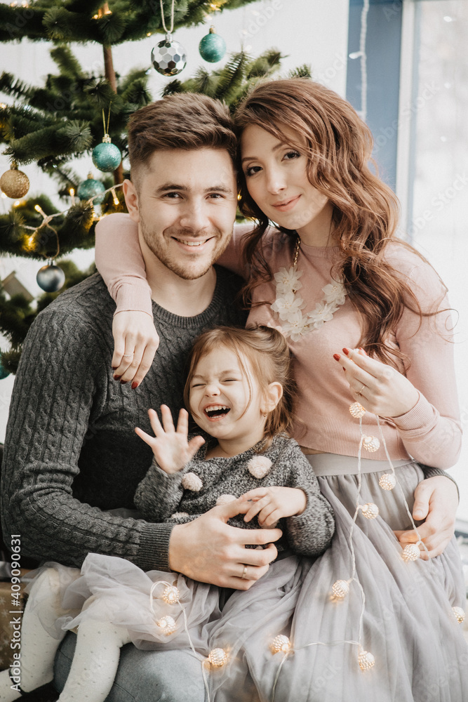 Young family with a daughter in festive outfits with a garland near the Christmas tree on New Years Eve Young family with a daughter in festive outfits with a garland near the Christmas tree on New Years Eve