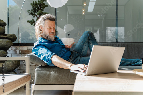 Mature man with a beard smiling and working with his laptop on a sofa photo