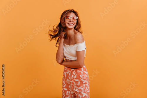 Tanned American girl with stylish haircut poses, looking at camera with smile. Portrait of joyful lady in summer outfit