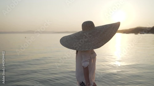 Middle shot of girl from the back in a big hat walking near the sea of Greece and turning her head to watch the sunset. Young woman wearing a big hat is walking on the coastline of Khalkidhiki island photo