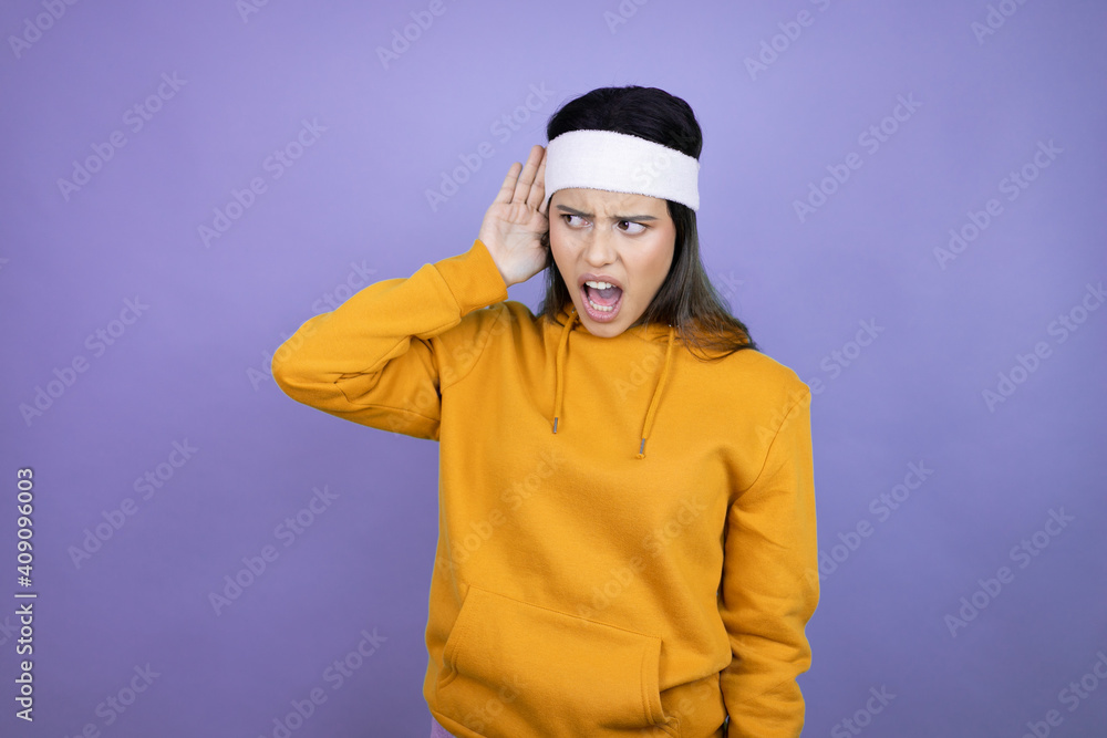 Young latin woman wearing sportswear over purple background surprised with hand over ear listening an hearing to rumor or gossip Young latin woman wearing sportswear over purple background surprised with hand over ear listening an hearing to rumor or gossip
