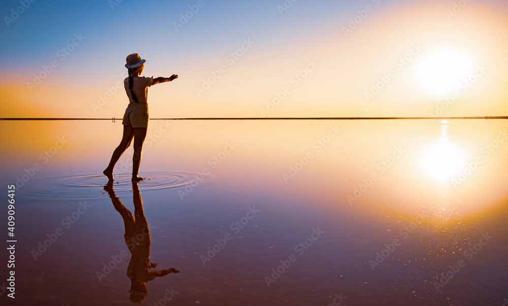 Fototapeta premium Lovely happy young girl walks and poses in a magical pose along the mirror pink salt lake enjoying the warm evening sun, looking at the fiery sunset and her reflection