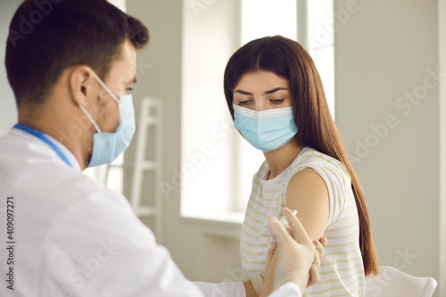 Young woman in medical face mask getting Covid-19 vaccine shot. Male nurse or doctor giving flu injection to female patient during seasonal vaccination campaign at clinic, hospital or health center