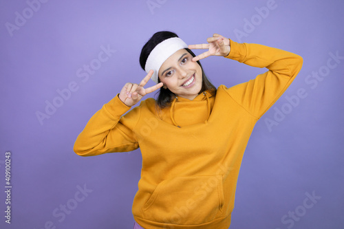 Young latin woman wearing sportswear over purple background Doing peace symbol with fingers over face, smiling cheerful showing victory