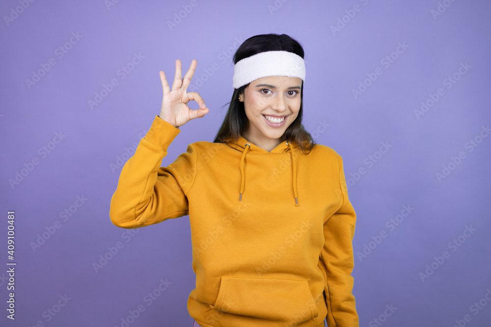 Young latin woman wearing sportswear over purple background doing ok sign with fingers and smiling, excellent symbol Young latin woman wearing sportswear over purple background doing ok sign with fingers and smiling, excellent symbol