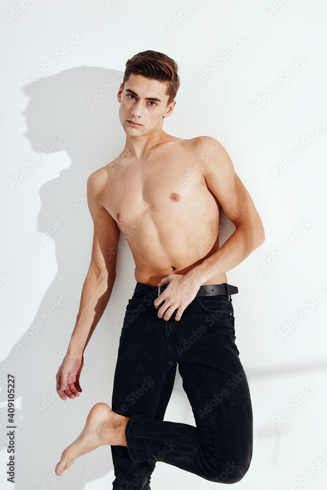 A half-naked guy in black trousers stands on one leg, leaning against the wall against a light background A half-naked guy in black trousers stands on one leg, leaning against the wall against a light background
