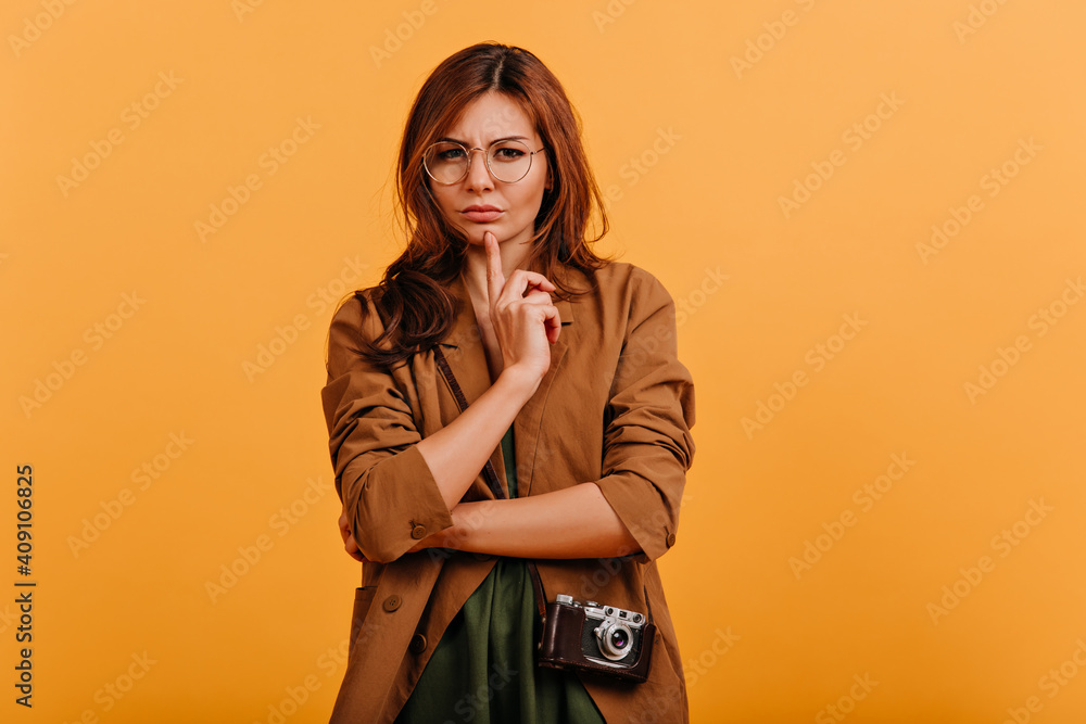 Pensive girl looking strictly at camera through glasses. Portrait of lady with curls in trench coat on isolated background Pensive girl looking strictly at camera through glasses. Portrait of lady with curls in trench coat on isolated background