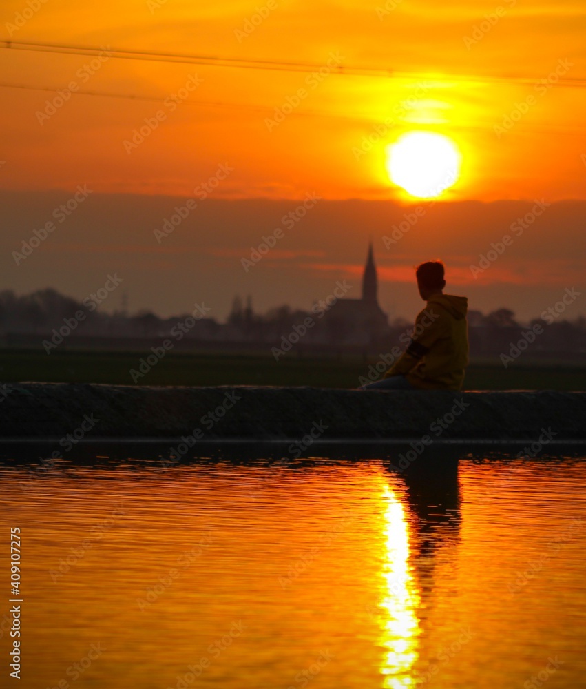 Fototapeta premium a amazing photo of a teenager sitting along the water, while the sun is setting. surrounded by farmersland, and a small town