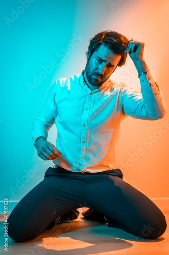 Lifestyle studio, a Caucasian man on his knees wearing a white shirt, illuminated with an orange and blue neon light