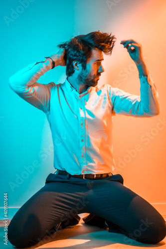 Lifestyle studio, a Caucasian man on his knees wearing a white shirt, illuminated with an orange and blue neon light